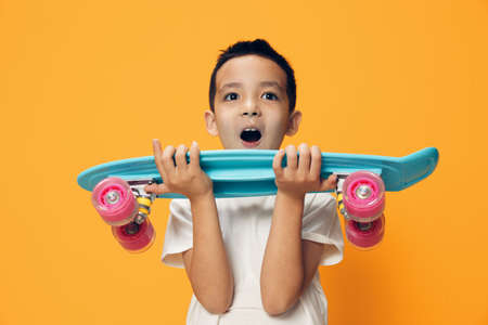 happy, little boy rejoices at the new skateboard in his hands standing on an orange background in a...