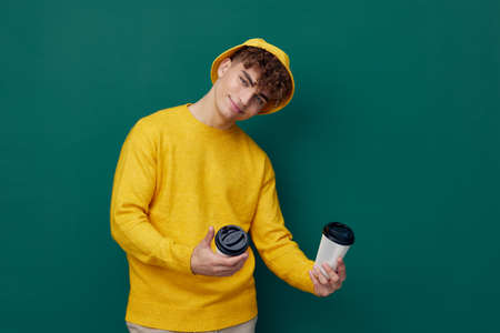 a joyful man stands on a green background in bright clothes and holds two coffee glasses in his hands, smiling affably at the cameraの写真素材