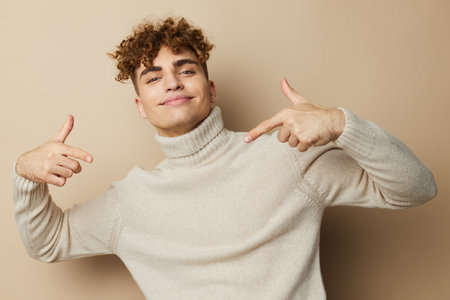 portrait of a handsome man with curly hair, standing on a beige background in a light turtleneck, pointing fingers at himself. A photo with an empty space for inserting an advertising layoutの写真素材