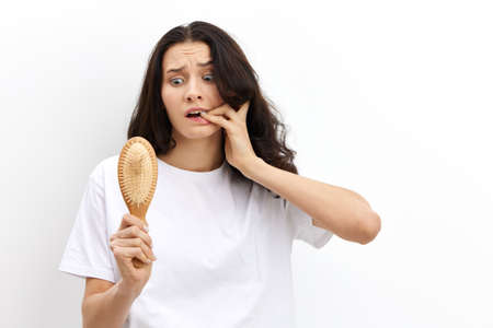 a sad, upset, puzzled woman with long black hair stands in a white T-shirt on a light background and holding her fingers to her mouth looks at her wooden combの写真素材