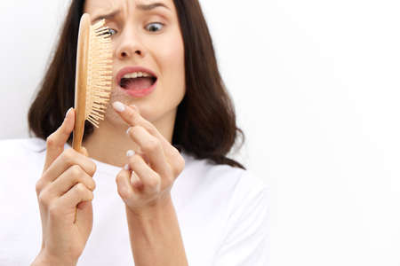 a close horizontal photo of an anxious, sad woman cleaning a comb from accumulated fallen hair and looking at them in panicの写真素材