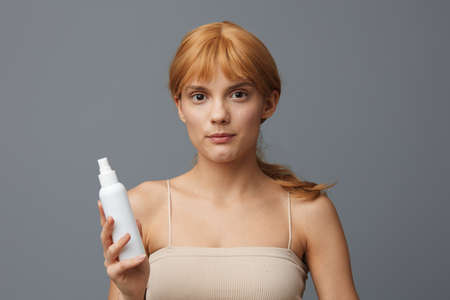 a woman in a white T-shirt with fresh clean skin, with red wavy hair gathered in a tail, he stands on a gray background with a white bottle in his hands.Horizontal studio shot.の写真素材