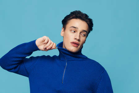 man is standing in blue zip-up sweater pulling the collar aside and looking away with a friendly...