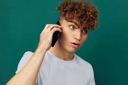 a funny, surprised man with curly hair stands against a green background and holds his black smartphone in his hand, talking on it, his mouth wide open in surpriseの写真素材