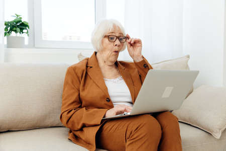 an attentive elderly woman in a brown suit is sitting at a laptop working remotely on the couch and peering at the text on the monitor holding her glasses on her faceの写真素材
