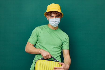 a handsome man stands on a green background, dressed in a yellow panama hat and with a mask on his face, holding a bright travel suitcase in his hand and looking at the cameraの写真素材