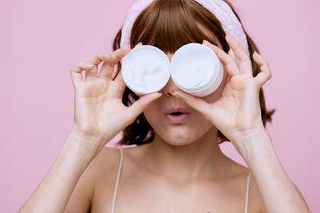 a funny woman stands on a pink background in a soft rim and holds a white jar of cream closing her eyesの写真素材