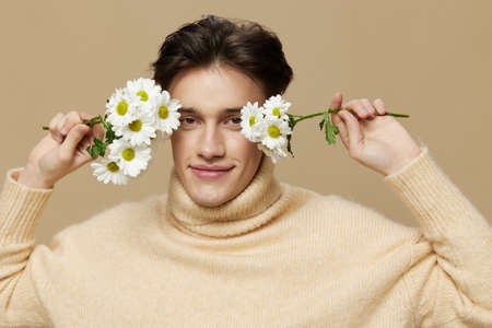 A gorgeousman with shining skin, with dark, short, short hair combed back, in a beige turtleneck with a high collar stands on a dark beige background with white daisies near his face.Close-up photo.の写真素材