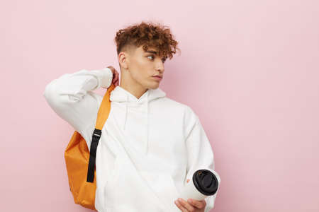 a nice man stands on a pink background in a white hoodie holding an orange backpack and a cardboard cup in his hands, looking pleasantly at the cameraの写真素材