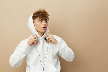 a cute, attractive man stands on a beige background in a white hoodie on his head and holds...