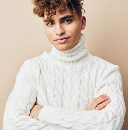 handsome, pleasant man with curly hair, standing in a white knitted sweater on a beige background and smiling pleasantly with his arms crossed on his chest. horizontal studio photo with an empty spaceの写真素材