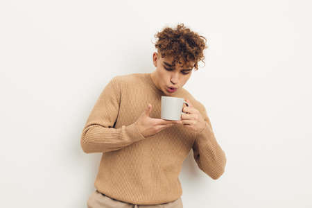 a cute, stylish, curly-haired man stands on a light background holding a white cup with a hot drink...