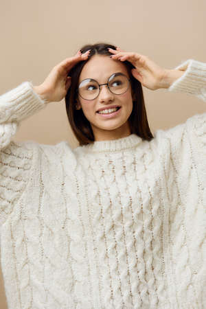a pretty woman stands on a light background in a white sweater and glasses, leaning her fingers...