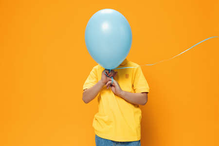 funny boy stands on a yellow background and holds a blue balloon in his hands, covering his face with itの写真素材