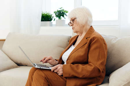 an attentive elderly woman with glasses is mastering new technologies using a laptop while sitting on the couch at home in a comfortable environment in a brown suitの写真素材