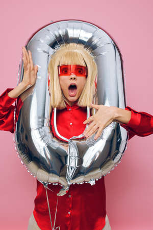 emotional woman standing on a pink background in a red shirt and glasses for a party, with a balloon...