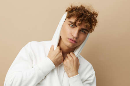 a stylish young man in a white hoodie, standing on a beige background and holding the hood on his...