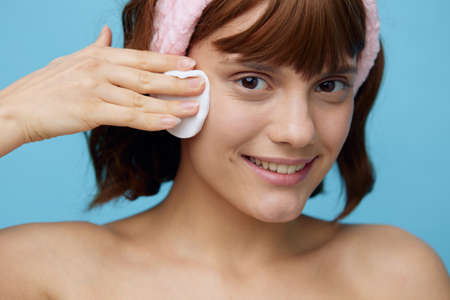 a girl with clean skin stands on a blue background in a white towel with a pink bandage on her head after a bath, removes makeup from her face with a cotton pad. Horizontal studio photography.の写真素材
