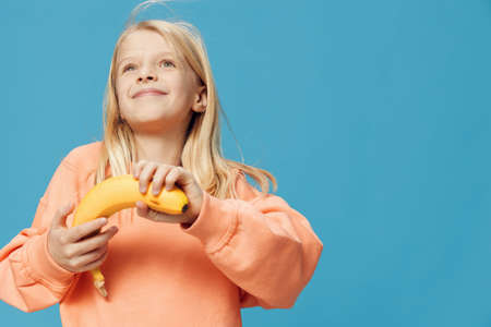 happy joyful girl posing fooling around with a banana in her hand and emotionally having fun posing. Horizontal photo in the studio on a blue background with empty spaceの写真素材