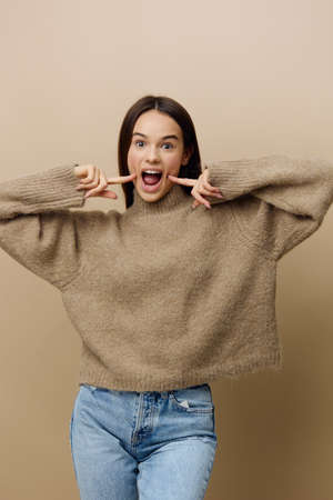 photo of a happy, joyful woman on a beige background looking emotionally into the camera and holding her index fingers on her cheeksの写真素材