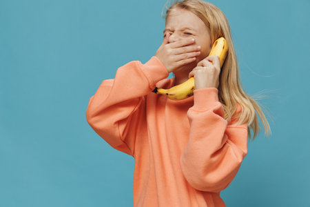 happy girl in bright orange clothes stands holding a banana in her hand near her ear and closes her...