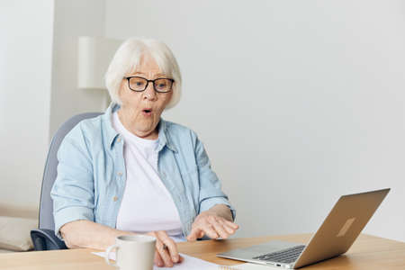 funny, emotional old lady with gray hair works from home on a laptop at home makes a funny facial expression looking at the monitorの写真素材