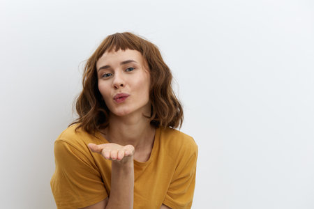 beautiful, attractive, redhead, romantic a woman poses standing in a yellow T-shirt on a white background and blows a kiss to the cameraの写真素材