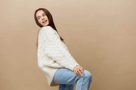 an elegant woman stands on a light background in a white sweater posing standing on one leg, and lifting the other slightly bending at the shin, bringing the body to the side to maintain balanceの写真素材
