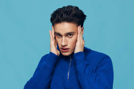 pleasant man stands in a blue zip-up sweater with his hands near his head touching his temples. Studio photo on a light blue background with an empty space for inserting an advertising layoutの写真素材