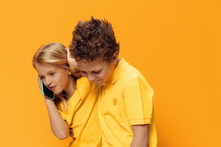 cute funny school-age children stand on a yellow background in bright clothes and hugging each other talking on the phoneの写真素材