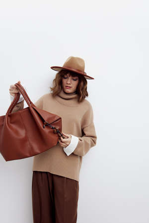 a sweet beautiful woman stands on a white background in a sweater, shirt and hat, looking at the bag with interest, holding it by the handle with one hand, holding it by the bottom with the other handの写真素材