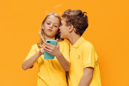 cute, funny children, school age, brother and sister stand on a yellow background and look into their smartphoneの写真素材