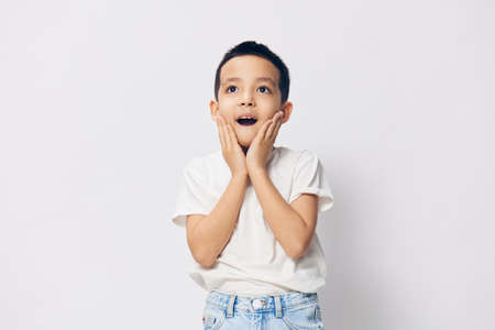 a cute little preschool-age boy stands on a white background in a white T-shirt, looks in surprise at the camera with his eyes wide open, raising his hands to his chin and opening his mouthの写真素材