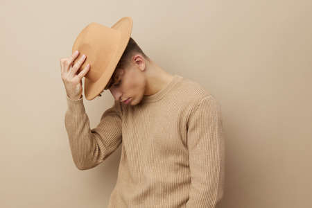 a handsome, young man stands on a beige background in a stylish sweater and holds a wide-brimmed hat in his hand, putting it on his head.の写真素材