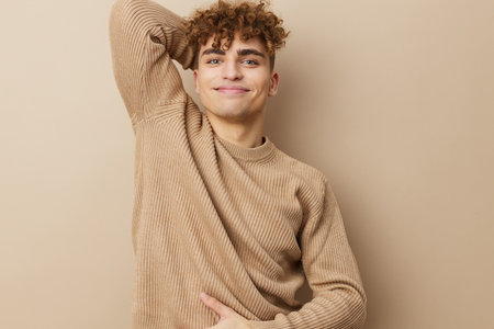 a charming, handsome man stands on a beige background in a sweatshirt and looks at the camera with a relaxed look, holding his hands on his curly hair, making a funny facial expressionの写真素材