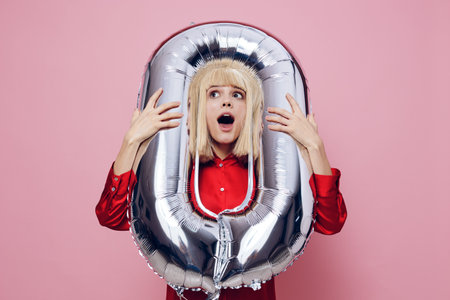 horizontal photo on a pink background of a beautiful, funny woman in a red shirt, wearing an inflatable ball in the form of the number zero around her neck and looking into the cameraの写真素材
