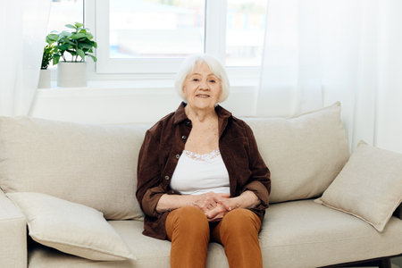 portrait of a pleasant elderly woman with a kind smile sitting on the sofa at home in a brown shirt smiling sweetly at the cameraの写真素材