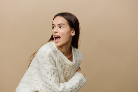 a perky woman is standing sideways on a light brown background in a white sweater slightly bent down, turning her head to the side and screaming, pressing her hand to herselfの写真素材