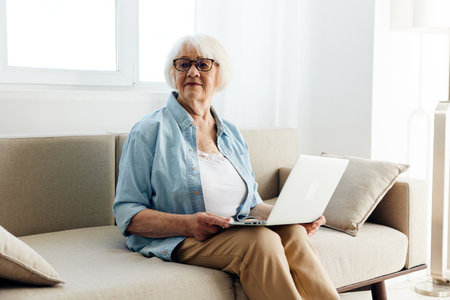 a happy, joyful, aged woman is sitting in a beautiful bright apartment with a laptop on a cozy beige sofa and looks with a pleasant smile on her faceの写真素材