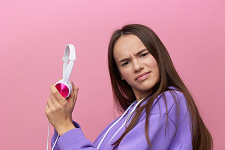 horizontal portrait of a sad, upset woman standing on a pink background and holding white headphones...