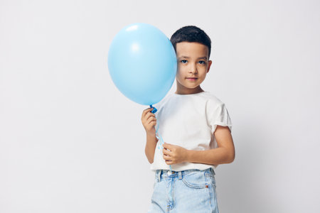 a cute, happy boy stands on a light background in a white t-shirt with a blue balloon in his hand and looks at the camera. Horizontal photo with blank space to insert advertising mockupの写真素材