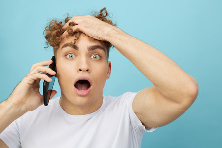 a funny, shocked man with curly hair stands on a blue background and holds his smartphone talking on it with his mouth wide open and clutching his head.の写真素材