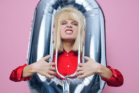 a funny, emotional woman stands on a pink background in a red shirt with an inflatable ball in the form of a zero and sticking her head into the hole makes a funny face showing her teethの写真素材