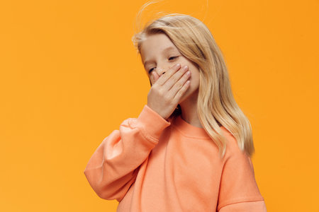 a beautiful girl, blonde, of school age, stands on an orange background in bright clothes and covers her mouth with her hand. Horizontal studio photography with blank space for advertising mockupの写真素材