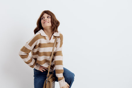 a pretty sweet woman stands on a light background in a striped sweater and a bag on her shoulder, put her hand on her hip and lifted one leg bending it at the kneeの写真素材