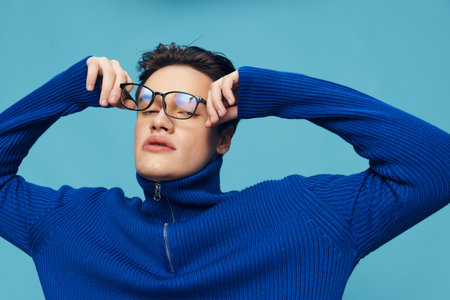 a handsome, attractive man stands on a light blue background in a blue zip-up sweater and holding his black-rimmed eyeglasses with his hands looks at the cameraの写真素材