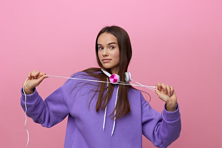 a happy, attractive woman stands with white headphones around her neck and plays with a wire from...