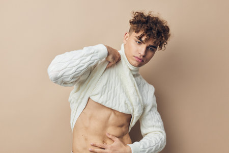 close horizontal photo of a handsome, attractive man standing in a white knitted sweater on a beige background, smiling pleasantly at the camera and showing his abs by lifting the edge of his clothesの写真素材