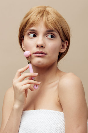 a close photo of a pretty woman on a beige background, standing with a facial massager in her hand, looking pleasantly into the cameraの写真素材