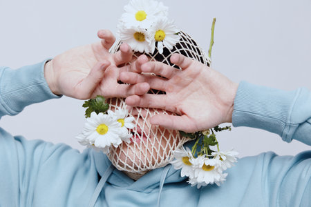 portrait of a strange man standing on a light background in a blue hoodie with a mesh on his face, to which white daisies are attached and poses relaxed with his hands covering his eyesの写真素材